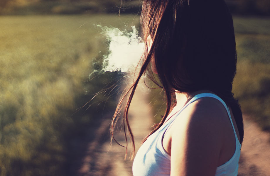 brunette woman standing in field outside facing away and blowing smoke out of mouth