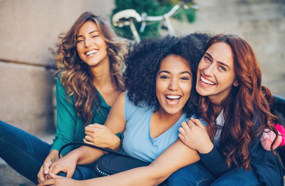 Multicultural group of young women sitting on steps outside in front of a bicycle smiling, hugging, and laughing