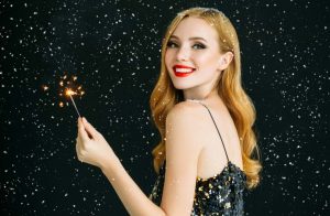 Blonde woman in black dress standing in front of a black background holding a lit sparkler.