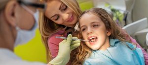 Young blonde girl with her mom sitting in the dental chair while a dentist uses a mirror to perform a dental exam.
