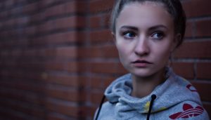 Blonde young woman wearing gray Adidas sweatshirt stands anxiously against a red brick wall wondering about her inflamed gums