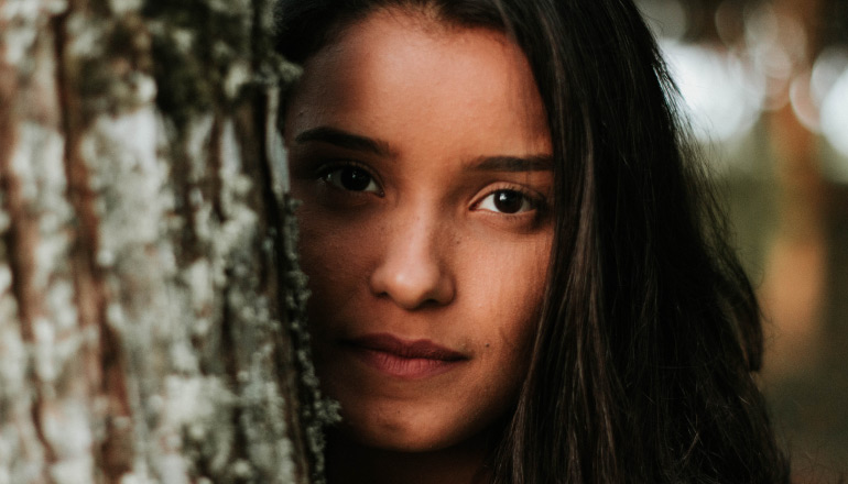 Brunette young woman hides part of her face behind the trunk of a tree due to worry over some acne