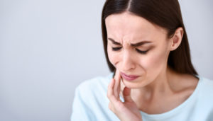 Brunette woman wearing a light blue shirt cringes and touches her cheek due to pain from sensitive teeth