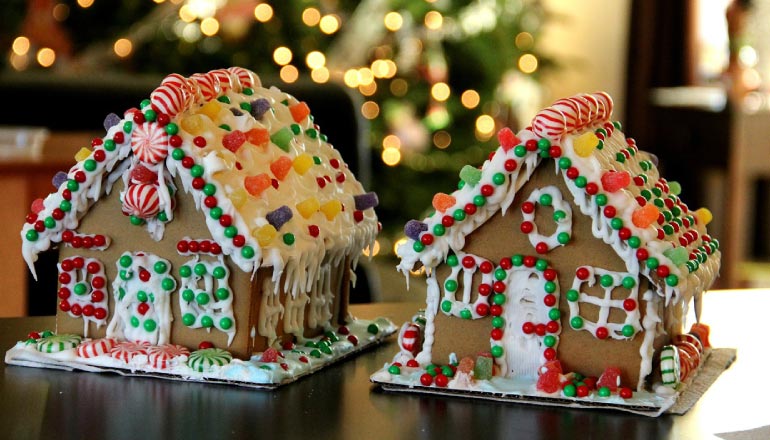 Two side by side gingerbread houses covered in sugary candies with a lit Christmas tree in the background